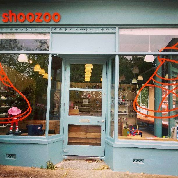 Smokey blue painted store front, with large windows either side of a central door.  Giant orange graphic shoe outlines adorn each window, and the shoozoo logo in orang raised lettering, all lower case, clean modern font, sits atop the fascia on the left.
